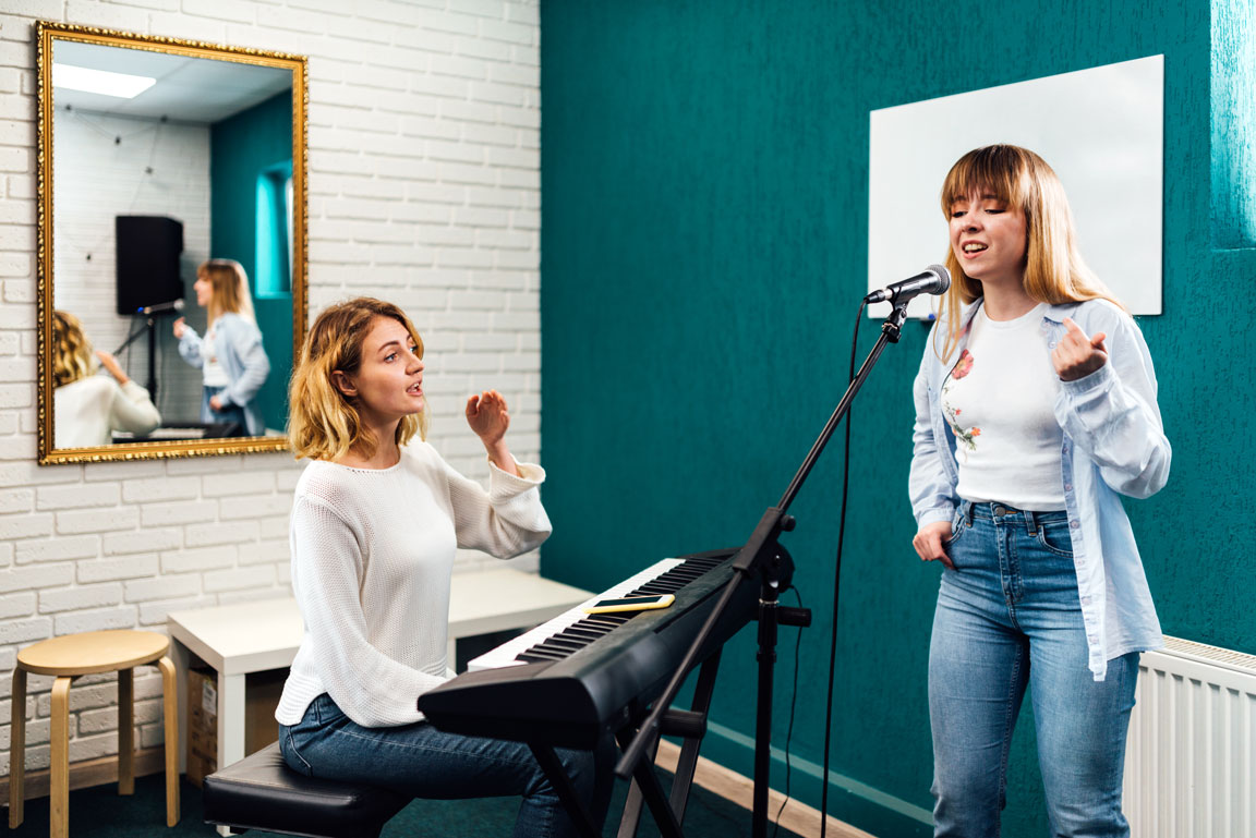 young adult female taking voice lessons while female music teacher plays keyboard